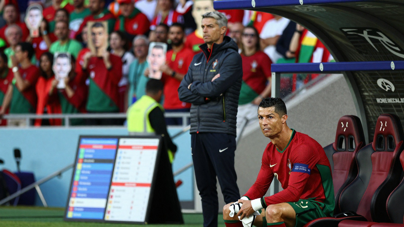 Jogador de futebol Cristiano Ronaldo sentado no banco com treinador e torcida ao fundo.